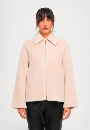 Woman with dark hair wears a pale pink fuzzy zip-up jacket with wide sleeves and black pants, standing against a plain white background.