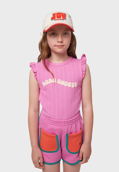 Top de tirantes rosa con mangas rizadas, con "ROBO CHOSES" en color crema. Pantalones cortos rosas con bolsillos naranjas y ribete en azul verdoso. Gorra blanca con logo rojo.