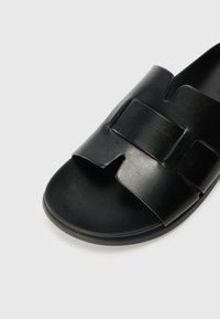 Black slide sandal with a thick, flat sole. Features a wide strap with a cut-out detail and smooth leather texture. Minimalist design.