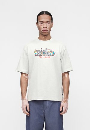 Νέος άνδρας με λεύκη στέκεται προς τα εμπρός φορώντας λευκό T-shirt New Balance με καρτούν χαρακτήρες και μπλε παντελόνι σε λευκό φόντο.