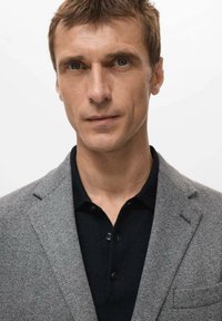 Szary blezer w jodełkę na czarnej koszulce polo. Blezer ma klapy z wycięciami i kieszeń, a na koszulce widoczne są guziki.