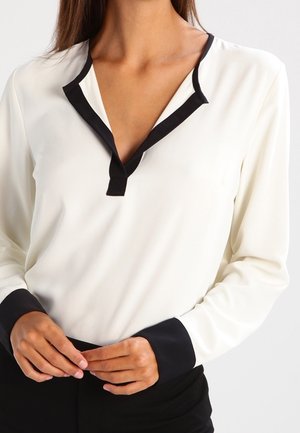 Femme portant un chemisier blanc avec un bord noir au col et aux poignets, mains doucement jointes à la taille.