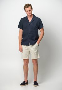 Camicia di lino azzurra a maniche corte con colletto e taschino frontale, abbinata a pantaloni corti crème. Il calzature presenta scarpette slip-on scure con suole intrecciate.