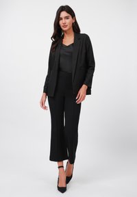 Blazer noir avec une ouverture à l'avant, deux poches, associé à un haut en satin noir et un pantalon ajusté, avec un ourlet court et des talons noirs pointus.