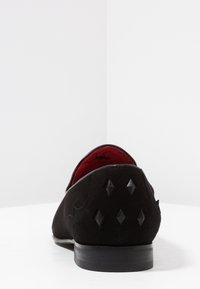 Zwarte suède schoen met een ronde neus, gewatteerd detail en zwarte studs op de hak. Rode voering. Platte zool.