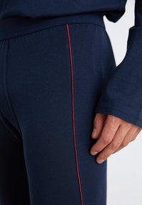 Marinefarbene Jogginghosen aus dehnbarem Material mit slim fit, ausgestattet mit einem roten Seitenstreifen und einem nahtlosen Bund. Hand liegt auf dem Stoff.