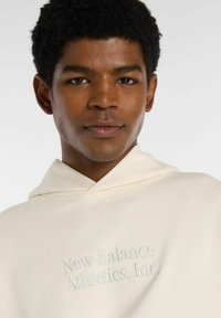 Joven con cabello corto y rizado que lleva una sudadera color crema bordada con "New Balance Athletics, Inc." en el pecho.