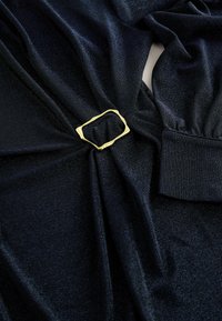Czarna, lśniąca tkanina o teksturowanym wykończeniu, z prostokątną, złotą klamrą na środku i mankietem przy rękawie.