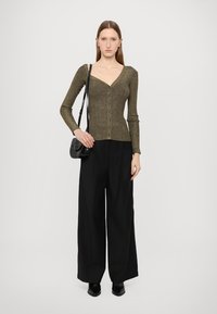 Olijfgroene metallic gebreide top met lange mouwen en een v-hals en knoopsluiting, gecombineerd met zwarte broek met wijde pijpen en een zwarte schoudertas.