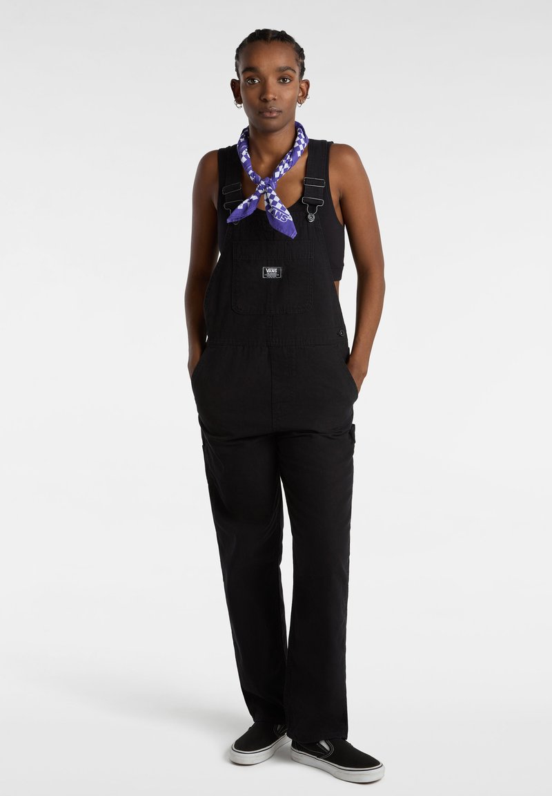 Zwarte overall gemaakt van duurzaam materiaal, met een voorzak, verstelbare banden en een relaxed fit. Gecombineerd met een paarse bandana en zwarte sneakers.