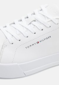 Zapatilla de cuero blanco con cordones planos, suela de goma y logotipo bordado "TOMMY HILFIGER". Textura suave y diseño de punta redonda.
