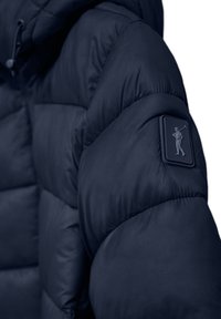 Marinblå pufferjacka med ett kviltat mönster, som har en texturerad tyg och en framträdande logopatch på vänster ärm.