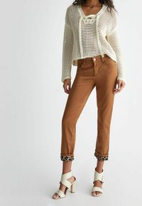 Kobieta ubrana w kremowy sweter o otwartym splotach, brązowe spodnie z podwiniętymi nogawkami i leopardzim wzorem wewnątrz, oraz kremowe sandały na wysokim obcasie z paskami na kostce.