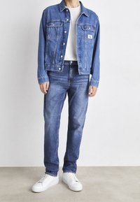 Denim jacket in medium blue with two chest pockets and button closure, paired with matching blue jeans and white sneakers. Smooth fabric texture.