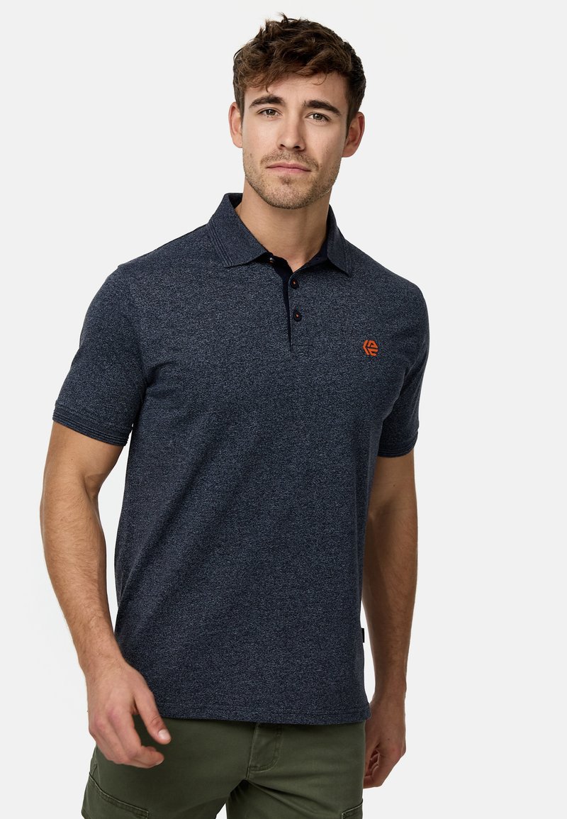 Marineblå polo skjorte med korte ærmer, struktureret stof, to-knap lukning, og et orange logo på venstre bryst. Sammen med grønne bukser.