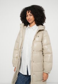 Casaco acolchoado bege com capuz, com design matelassê e fechos de pressão. Usado por cima de uma camisa branca às riscas e jeans azuis de ganga.