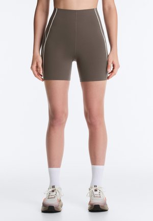 Personne portant un short cycliste marron taille haute avec des coutures blanches, des chaussettes blanches et des chaussures de course beiges, debout sur fond blanc.