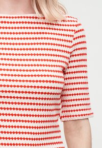 Εγγύς πλάνο ενός υφασμάτινου λευκού τοπ με οριζόντια κόκκινα διαμάντια και μισά μανίκια, σε άτομο με ανοιχτόχρωμο δέρμα και ξανθά μαλλιά.