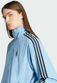 Lichtblauwe zip-up jas met zwart drie-strepen ontwerp langs de schouders, gemaakt van lichtgewicht materiaal. Adidas-logo op de linkerborst.