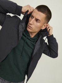 Czarna kurtka zapinana na zamek o gładkiej fakturze, z kapturem, noszona na ciemnozielonym hoodie. Model poprawia kurtkę z skoncentrowanym wyrazem twarzy.