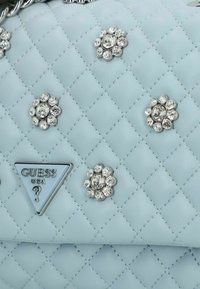 Borsa a tracolla azzurro chiaro trapuntata con decorazioni floreali in cristallo e logo triangolare di Guess. Caratterizzata da hardware argentato e una texture liscia.