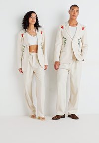 Costum crem cu broderie în formă de inimi roșii și modele de șerpi verzi. Pantaloni largi, asociați cu un top alb scurt și încălțăminte mixtă.