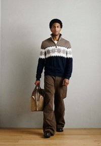 Camisola de malha com padrão castanho e azul-marinho, gola com fecho de correr e padrão de flocos de neve brancos; combinada com calças castanhas de perna larga e uma mala tipo duffel em tom bege.