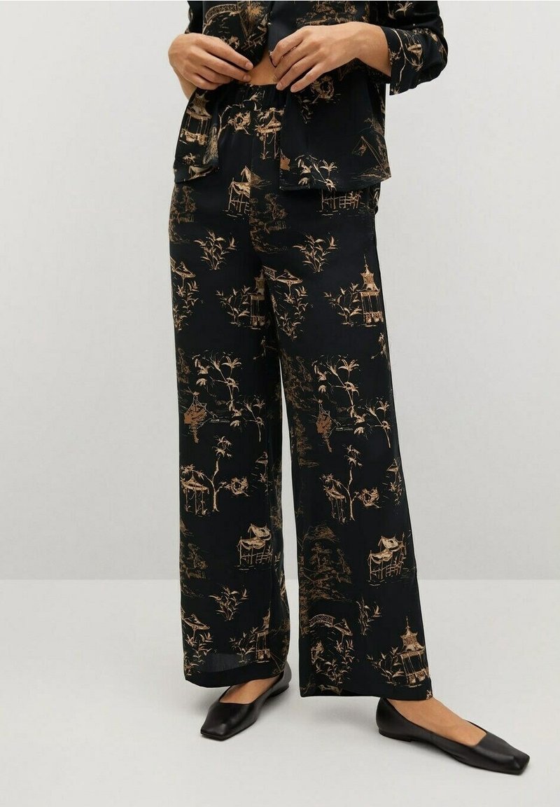 Femme portant un pantalon noir à jambes larges avec un imprimé beige asiatique de pagode et de bambou, associé à des ballerines noires à bout carré.