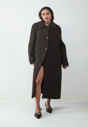 Brauner, strukturierter, langer Mantel mit breitem Kragen, Knopfverschluss und seitlichem Schlitz, kombiniert mit schwarzen spitzen Schuhen. Weißer Hintergrund.