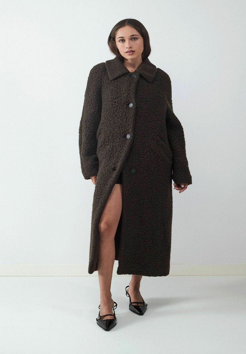 Bruine, gestructureerde lange jas met een brede kraag, knoopsluiting en zijsplit, gecombineerd met zwarte puntige schoenen. Witte achtergrond.