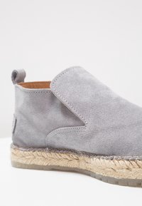 Gros plan d'une chaussure grise en daim sans lacets avec une semelle en corde tressée et une languette à l'arrière.