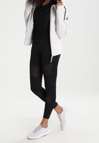 Leggings sportivi neri con pannelli in rete, abbinati a una maglietta nera e a una giacca bianca con zip. Sneakers bianche completano l'outfit.