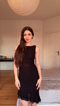 Νέα γυναίκα με μακριά καστανά μαλλιά που φορά αμάνικο μαύρο φόρεμα στέκεται σε ένα υπνοδωμάτιο με ξύλινο πάτωμα και λευκές πόρτες.