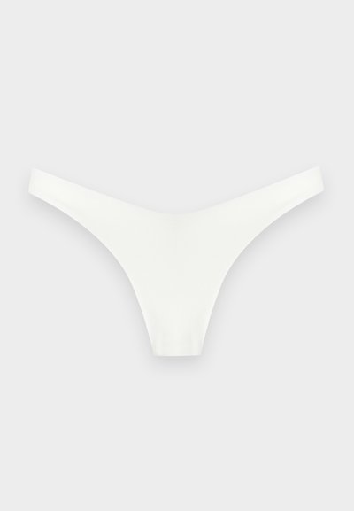 Sous-vêtement blanc sans couture de type string, posé à plat sur un fond gris clair, montrant les bretelles avant et latérales.