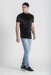 Čierne sametové tričko s krátkym rukávom a hladkou textúrou, nosené s bledomodrými roztrhnutými džínami a čiernymi teniskami. Model stojí v profilu.