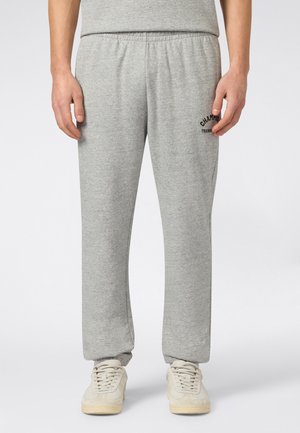 Mężczyzna w jasnoszarych spodniach dresowych z elastycznym pasem i czarnym napisem "Champion Training" na górnej części uda, w połączeniu z kremowymi trampkami.