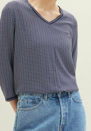 Persoon draagt een marineblauwe top met patroon en V-hals met driekwart mouwen, gestopt in lichtblauwe hoge taille jeans.