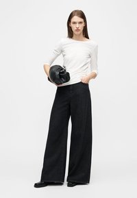 Γυναίκα που φοράει λευκό μακρυμάνικο τοπ, παντελόνι μαύρο με φαρδιά πόδια, μαύρα παπούτσια, κρατώντας μαύρη δερμάτινη τσάντα χειρός πάνω σε λευκό φόντο.