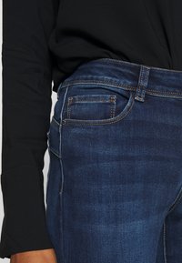 Jeans de mezclilla azul oscuro con un diseño ajustado, que cuentan con un bolsillo trasero y costuras de contraste en las costuras. Parte de una camiseta negra es visible.