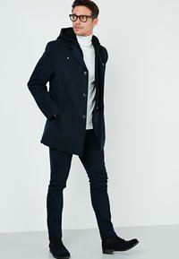 Abrigo de lana azul marino con capucha, botones en la parte delantera, bolsillos laterales, combinado con pantalones negros y botines, sobre un fondo blanco.