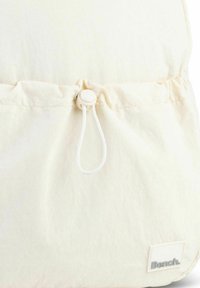 Cream fabric drawstring backpack with a smooth texture, featuring a plastic toggle closure and a white label with gray lettering at the bottom.