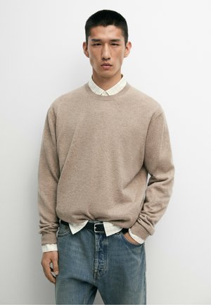 Homme portant un pull beige par-dessus une chemise blanche à carreaux et un jean bleu, debout avec une main dans la poche devant un fond blanc uni.