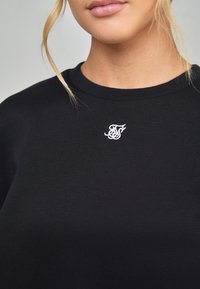Sudadera de algodón negra con cuello redondo. Presenta un pequeño logo bordado en blanco en el pecho. Textura suave, corte relajado.