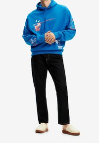 Modrá mikina s grafickými potisky, kombinovaná s černými džíny s kontrastním prošitím a krémovými dřeváky.