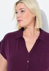 Shirt v barvě švestky s krátkým rukávem, která má zapínání na knoflíky, límeček a levou kapsu na hrudi s růžovými knoflíky a měkkou texturou látky.