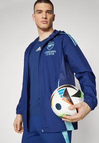 Giacca da calcio blu navy con dettagli turchesi, con il logo dell'Arsenal. Indossata sopra una maglietta blu scuro, mentre tiene un pallone da calcio multicolore.