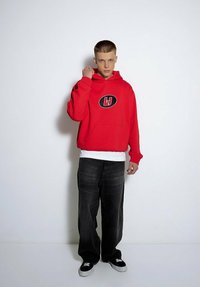 Czerwona bluza z kapturem z czarną owalną grafiką i kieszenią, noszona na białej koszulce. Uzupełniona luźnymi czarnymi dżinsami i czarnymi trampkami.