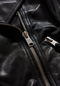 Musta nahkatakki, joka on varustettu näyttävällä metallivetoketjulla, jossa on teksturoitu vetotappi. Takki esittelee sileän pinnan ja tyylikkään muotoilun.