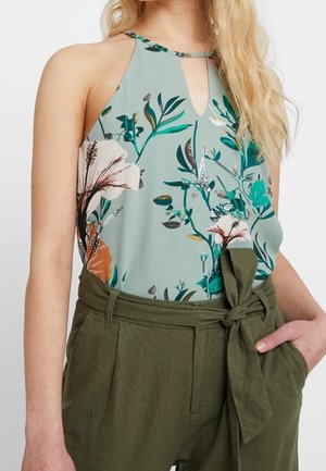 Vrouw draagt een lichtgroene bloemen haltertop en olijfgroene hoge taillebroek met een gestrikte riem, hand in zak.