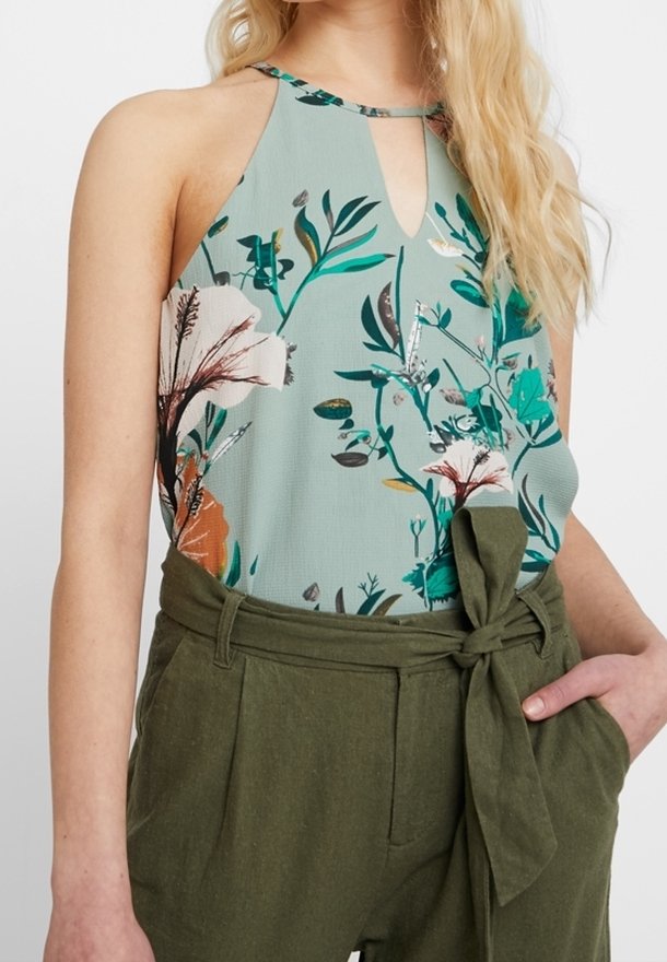Femme portant un haut dos nu à fleurs vert clair et un pantalon taille haute vert olive avec une ceinture nouée, main dans la poche.